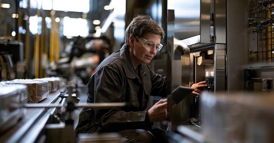 Field engineer in safety glasses checking a tablet beside an open machine panel on a busy, brightly lit production line