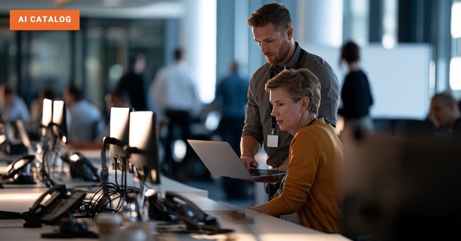 Employee holds out his laptop to an IT colleague at her desk in a busy open-plan office as they work through a problem